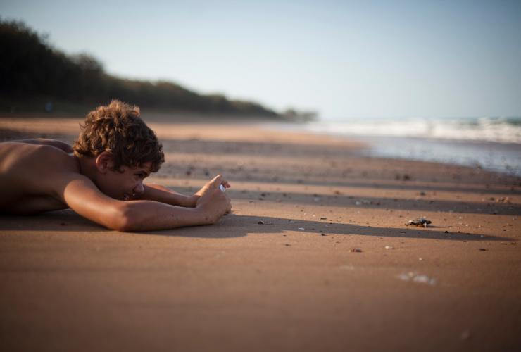 A child laying on the sand to take a photo of a baby turtle approaching the ocean at Mon Repos, Bundaberg, Queensland © Tourism and Events Queensland/Rowan Bestmann