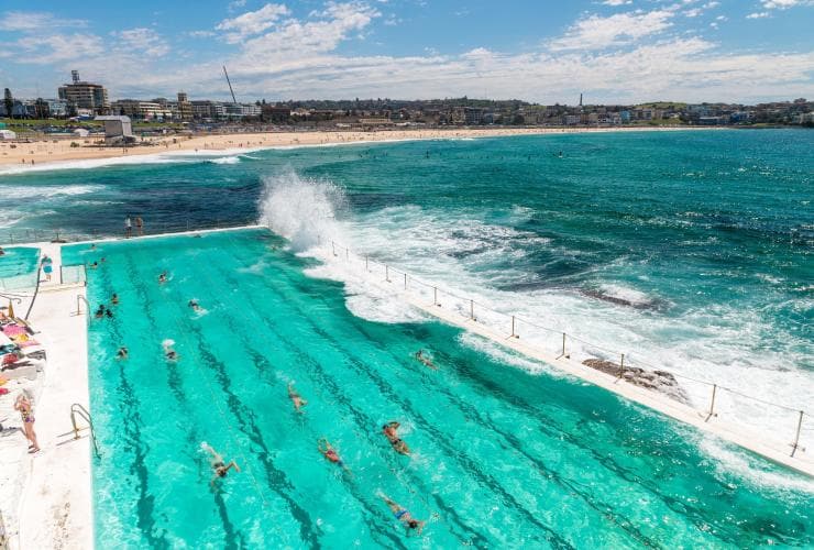 View over people swimming laps in the turquoise water of Bondi Icebergs Pool as waves crash into the pool, with Bondi beach in the background in Sydney, New South Wales © Destination NSW