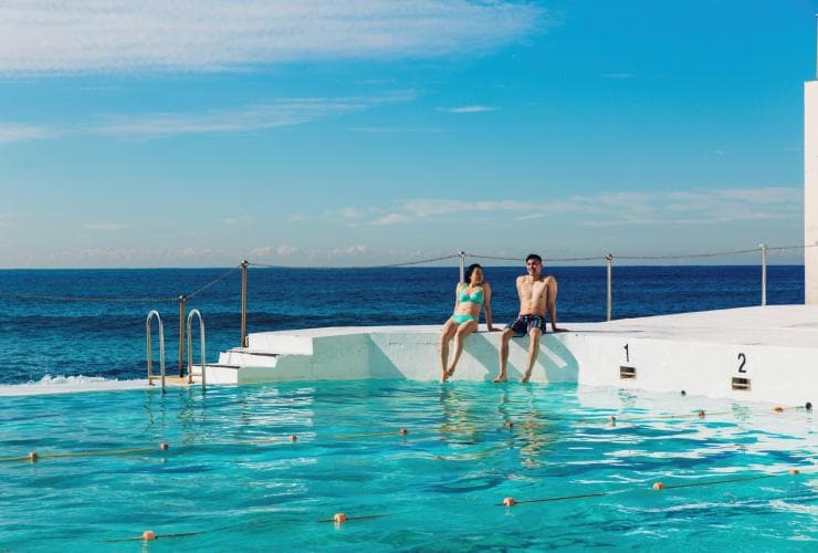 A man and woman sitting on the edge of Bondi Icebergs with their feet in the water and the ocean behind them in Sydney, New South Wales © Destination NSW