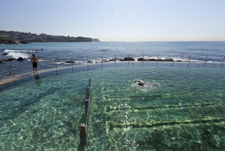 A person swimming in the sparkling water of Bronte Baths with another person standing on the edge overlooking the ocean at Bronte Beach, Sydney, New South Wales © Andrew Gregory/Destination NSW