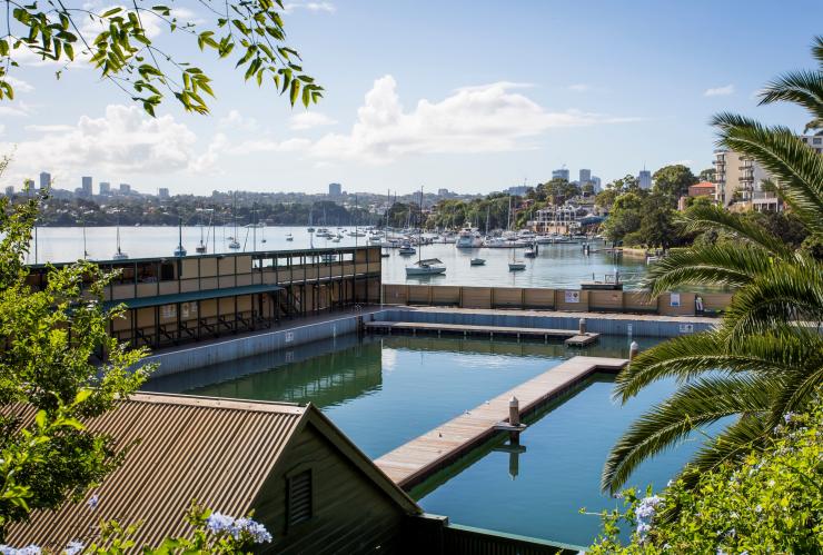 View through the trees into the pools of Dawn Fraser Baths with boats and waterfront buildings in the background in Balmain, Sydney, New South Wales © Destination NSW