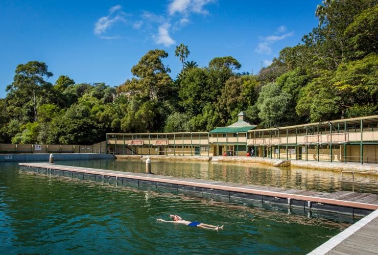 A person floating in the blue-green water of Dawn Fraser Baths with the building’s historic walls surrounded by trees in the background in Balmain, Sydney, New South Wales © Destination NSW