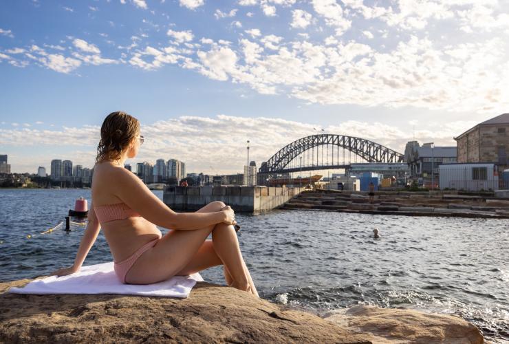 A woman sitting on a sandstone rock looking past Marrinawi Cove to the Sydney Harbour Bridge while people swim below her, Barangaroo Reserve, Sydney, New South Wales © Images for Business