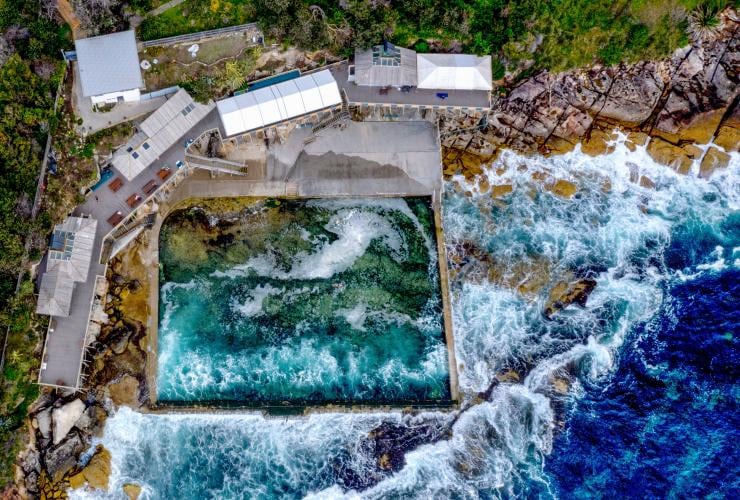 Aerial view over Wylies Baths ocean pool with people swimming and waves crashing over coastal rocks surrounding the pool, Coogee, New South Wales © Destination NSW