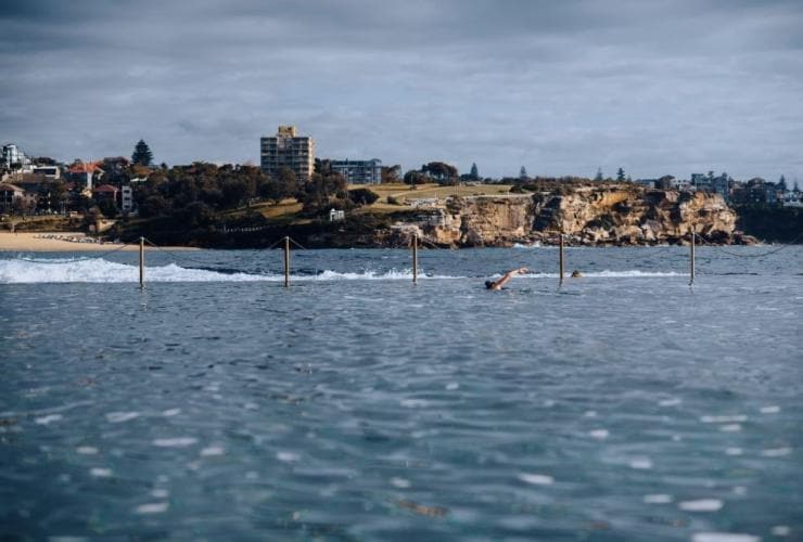 Close up of the blue water of Wylies Baths in the foreground with a person swimming in the background and a headland beyond in Coogee, New South Wales © Destination NSW