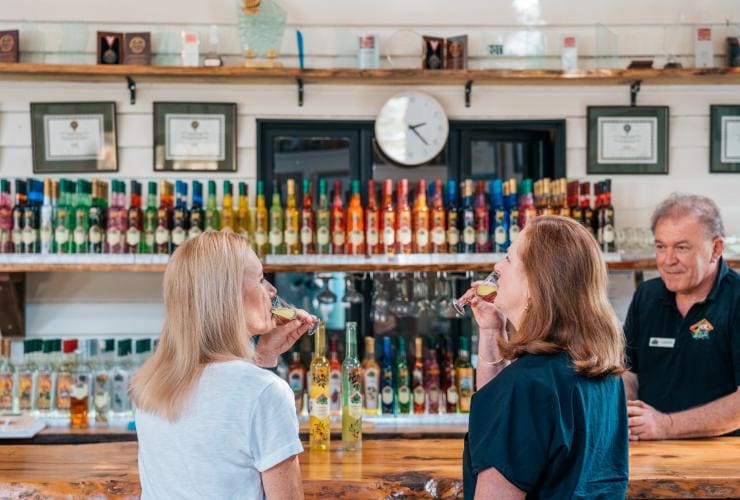 Deux femmes sirotant un verre de liqueur au bar, devant un barman et plusieurs bouteilles colorées de spiritueux exposées derrière le comptoir, lors d'une visite guidée avec Vino Bus à la Tamborine Mountain Distillery, Brisbane, Queensland © Tourism and Events Queensland