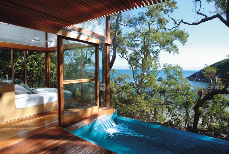 Une piscine à débordement près d'un lit situé dans une chambre entourée de baies vitrées, près d'arbres verdoyants dont le feuillage laisse entrevoir l'eau bleue de l'océan, au Bedarra Island Resort, Grande Barrière de Corail, Queensland © Bedarra Island