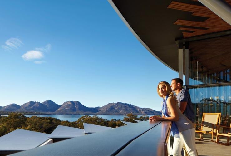 Un homme et une femme se penchant sur un balcon surplombant Coles Bay, avec le bush et les sommets des montagnes en arrière-plan, au Saffire Freycinet, Freycinet, Freycinet National Park, Tasmanie © Saffire Freycinet
