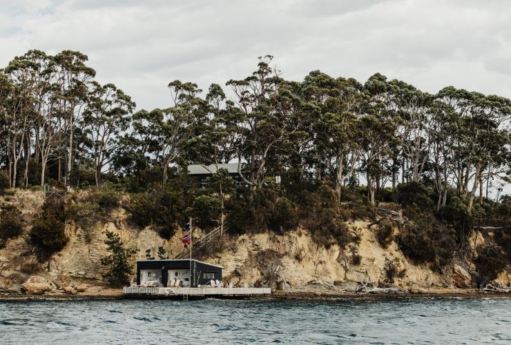 Vue depuis l'océan sur une eau bleue et tranquille, et sur un cottage de bord de mer situé sous de petites falaises rocheuses et des arbres anciens à Satellite Island, Tasmanie © Adam Gibson
