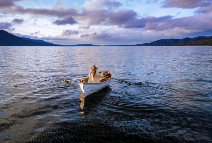 Une femme sur une barque, naviguant avec des rames sur une eau tranquille sous un ciel bleu et violet près de Satellite Island, Tasmanie © Adam Gibson