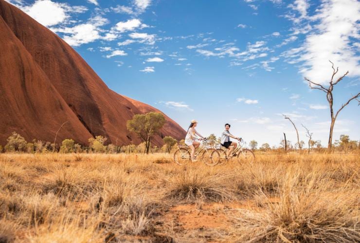 Dua orang bersepeda di sepanjang padang rumput dengan dinding batu merah Uluru menjulang tinggi di belakang mereka di Uluru-Kata Tjuta National Park, Northern Territory © Tourism NT