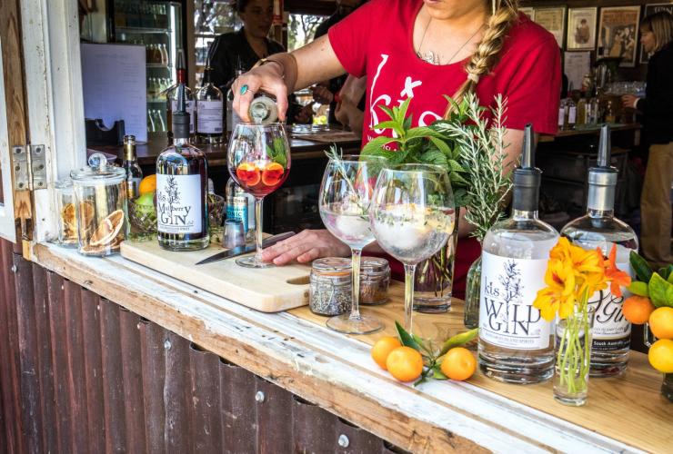 Foto dari jarak dekat seorang bartender menuang koktail buah di bar yang dikelilingi dengan buah-buahan segar, herba, dan beberapa botol gin di Kangaroo Island Spirits, Kangaroo Island, South Australia © South Australian Tourism Commission