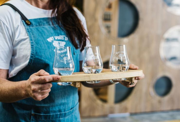 Foto dari jarak dekat seorang bartender memegang papan pencicipan berisi tiga minuman beralkohol yang berbeda di Manly Spirits Co., Manly, New South Wales © Alana Dimou