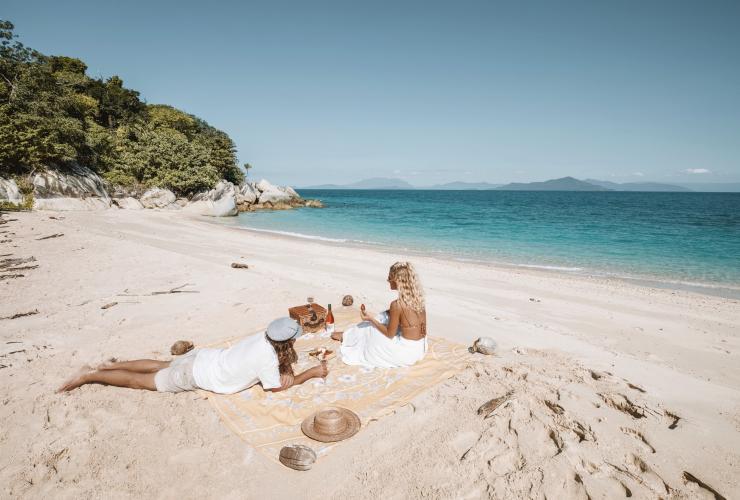 Dua orang sedang bersantai duduk di atas tikar piknik dengan minuman anggur, stroberi, dan keju di pantai berpasir putih di samping samudra biru tenang di Bedarra Island Resort, Great Barrier Reef, Queensland © Bedarra Island Resort/Elise Cook
