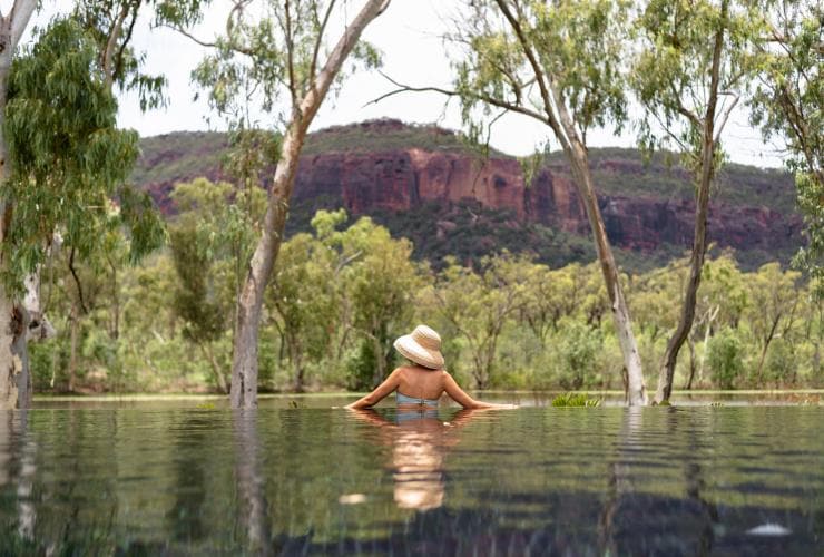 Seorang wanita berenang di kolam dengan air yang mencerminkan pepohonan dan pegunungan di sekelilingnya di Mt Mulligan Lodge, Mount Mulligan, Queensland © The Rambler Co
