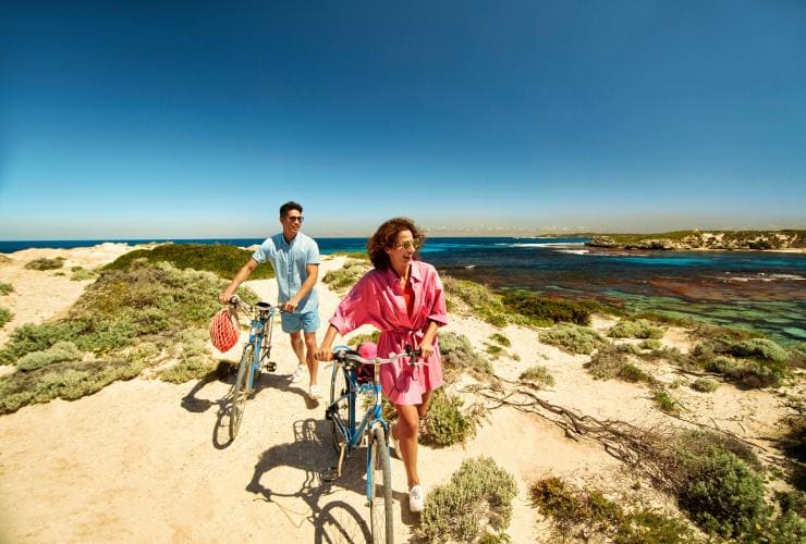 Due persone che spingono le biciclette lungo un sentiero sabbioso davanti al limpido oceano blu a Rottnest Island, Western Australia © Georges Antoni