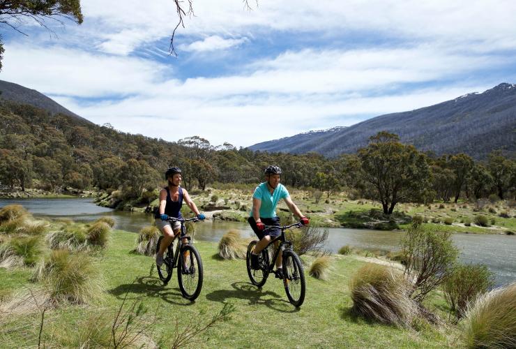 Due persone in bicicletta lungo un sentiero erboso accanto a un fiume circondato dal verde a The Diggings, Kosciuszko National Park, New South Wales © Destination NSW