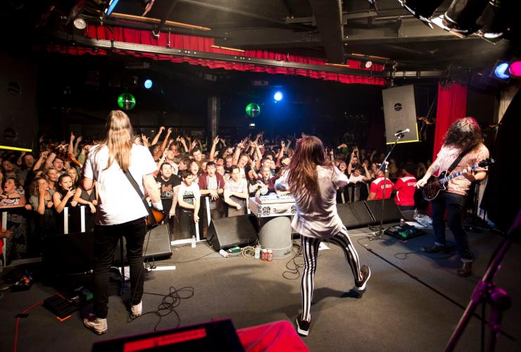 Vista di una band di spalle che si esibisce su un palco davanti a una folla di persone con le mani alzate al Corner Hotel, Richmond, Victoria © Zo Damage