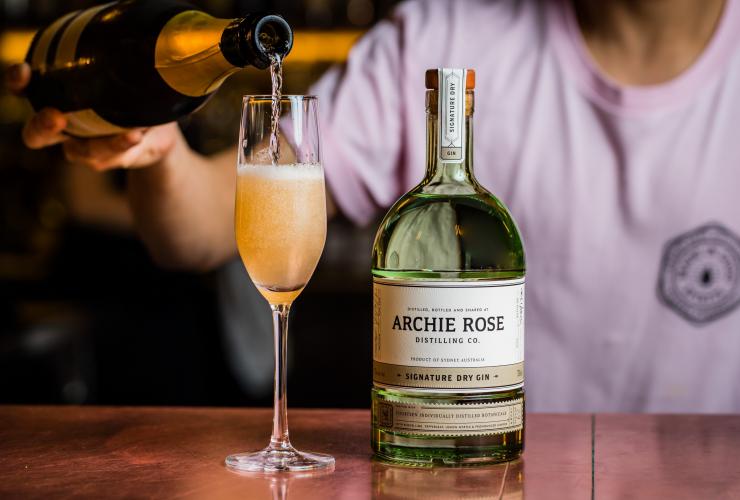 Primo piano di un barista che versa champagne in un calice accanto a cui si trova una bottiglia di Signature Dry Gin da Archie Rose, Sydney, New South Wales © Archie Rose