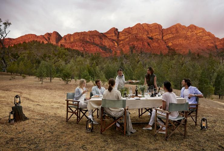 Gruppo di persone che cenano all'aperto in una pianura erbosa circondata dagli alberi, con le cime delle montagne illuminate di rosso in lontananza all'Arkaba Conservancy, Flinders Ranges, South Australia © Wild Bush Luxury
