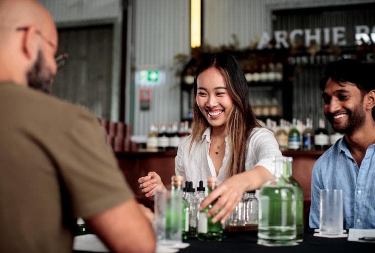 ニュー・サウス・ウェールズ州、シドニー、アーチー・ローズ・ディスティラリーにて、テーブルに並ぶジンの小瓶に笑顔で手を伸ばす女性と男性 © Destination NSW