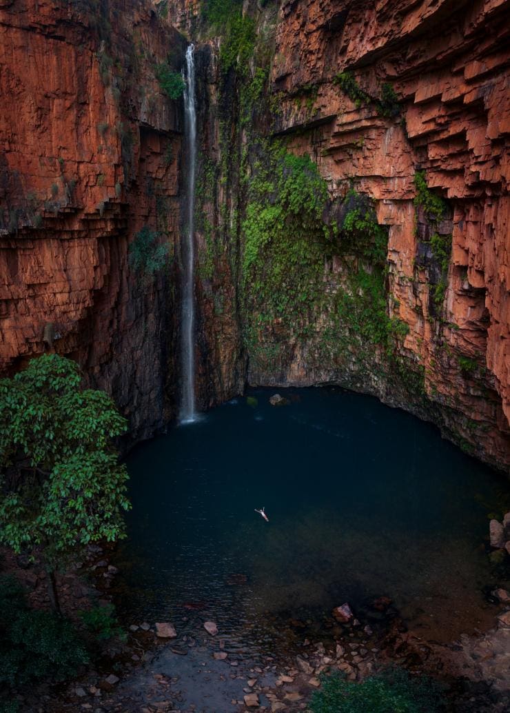 西オーストラリア州、エル・クエストロ・ウィルダネス・パーク、エマ渓谷にて、赤い岩の崖を伝って穏やかな滝が流れ込む深い青の淵で仰向けに浮かぶ人の姿を上方から © Tourism Australia