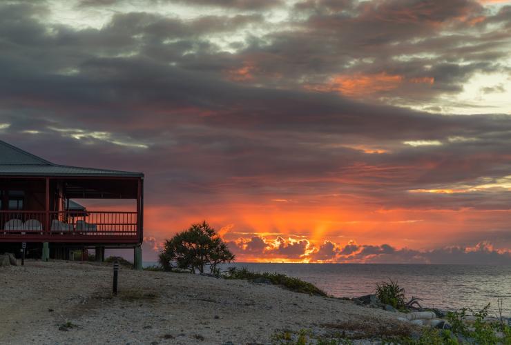 クイーンズランド州、ウィットサンデー諸島、キャンプ・アイランド・ロッジ、鮮やかなオレンジの日没をバックに立つ海辺の建物 © Camp Island Lodge
