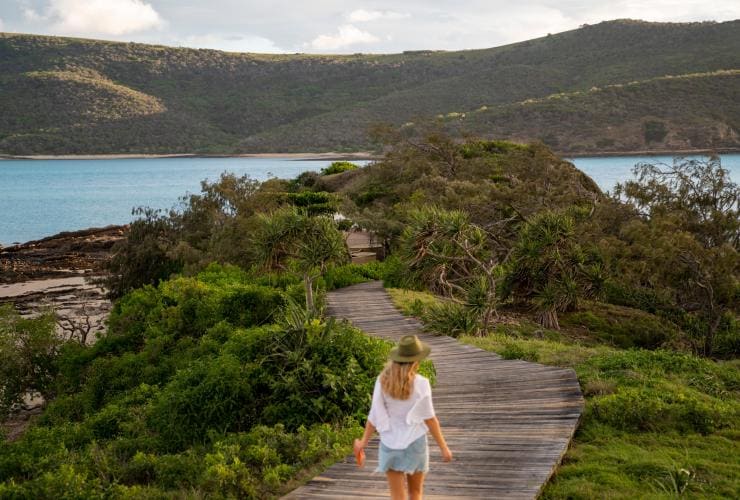 クイーンズランド州、サザン・グレート・バリア・リーフ、パンプキン島にて、遠くに海を見晴らす緑のブッシュランドに囲まれた木道を歩いていく女性 © Tourism and Events Queensland
