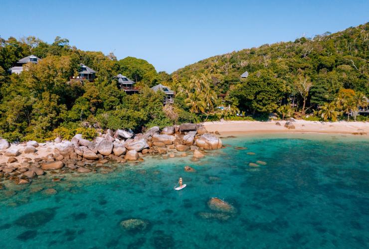 昆士蘭州大堡礁班達拉島度假村清澈碧藍的海洋上一人在岩石海岬附近白色沙灘旁玩站立划槳滑浪板，茂密綠林之中是度假村的別墅©James Vodicka