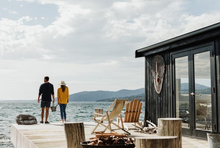 塔斯曼尼亞衛星島一對情侶站在海洋旁的度假小屋露台邊緣，露台擺放了躺椅和營火盆©Adam Gibson