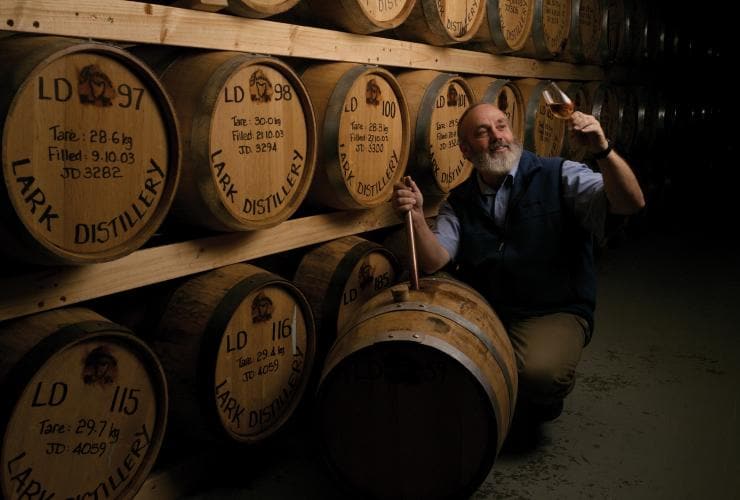 塔斯曼尼亞荷伯特Lark Distillery釀酒廠內一男子蹲在一排疊得高高的橡木酒桶旁，正在欣賞一杯剛倒岀的佳釀©Osborne Images